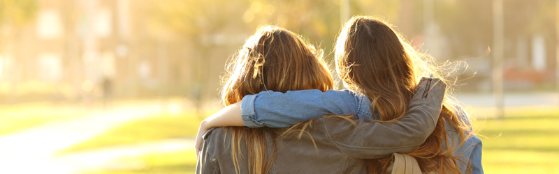 To unge jenter som holder rundt skuldrene til hverandre. Bildet er tatt utendørs i vårvær.